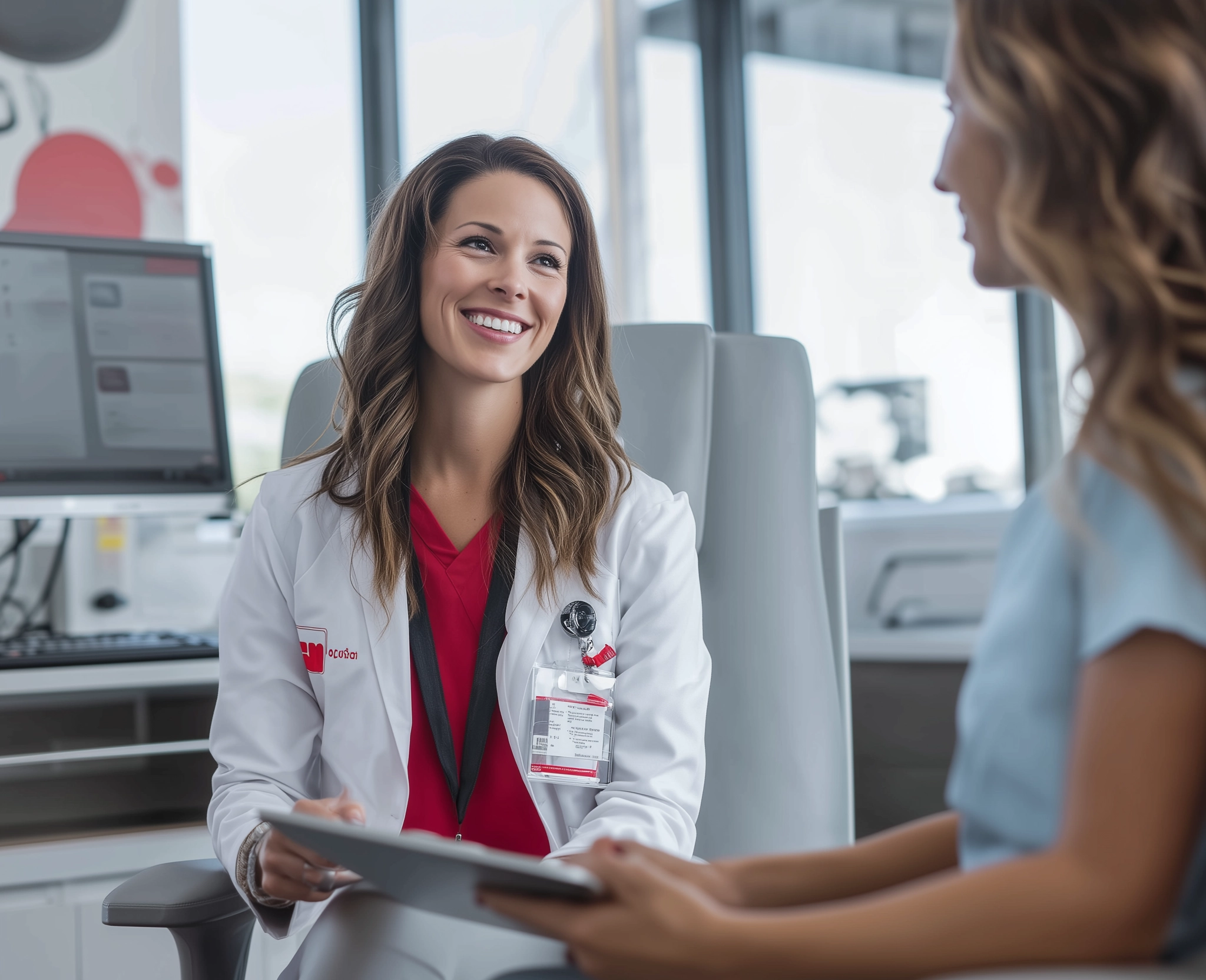 A clinician in a white coat smiles while reviewing a tablet with a patient in a bright medical office.